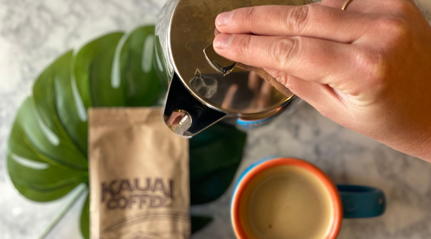 An overhead view of a hand pressing the plunger on a silver French Press. A bag of Kauai Coffee Estate Reserve is visible on the table