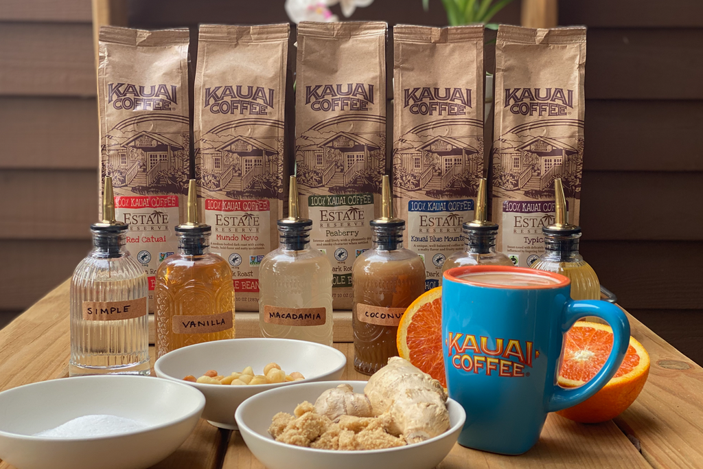 five bags of estate reserve kauai coffee with six home made coffee syrups sit on a wooden table. In the foreground are the syrup ingredients including sugar, macadamia nuts, ginger and a sliced orange. 