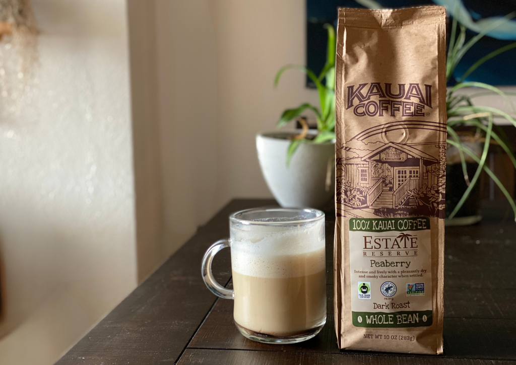 a bag of kauai coffee estate reserve peaberry dark roast sits on a table next to a breve coffee. some plants are visible in the background. 