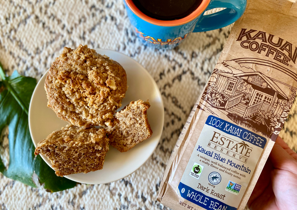 two coffee cake muffins sit on a white plate. A blue kauai coffee mug filled with black coffee and a bag of kauai blue mountain coffee sit on a white table cloth.