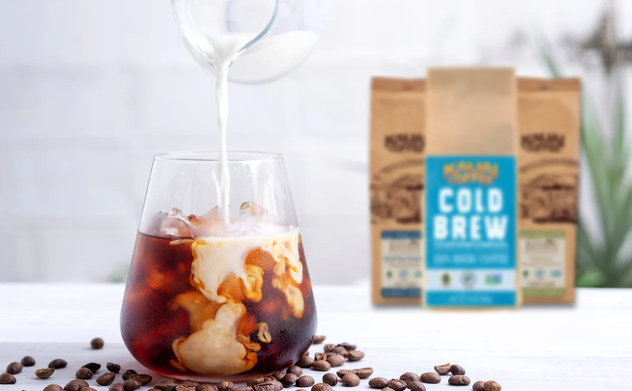 a short glass with cold brew coffee has milk poured in. Three bags of kauai coffee are visible in the background