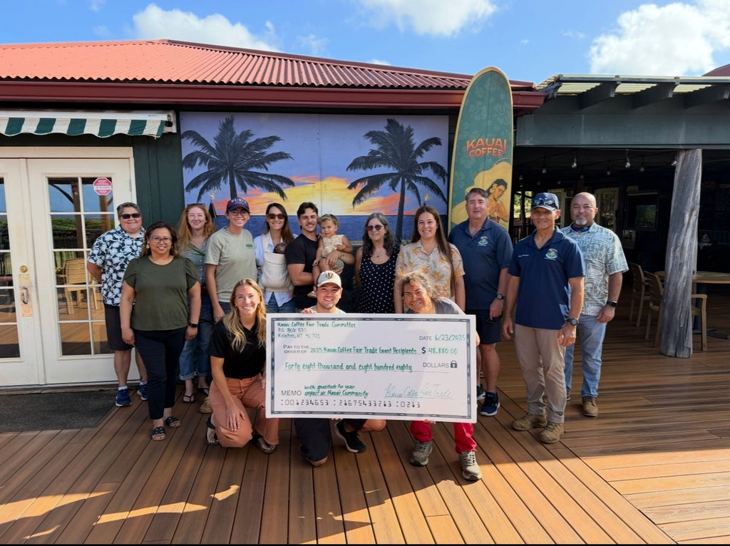 Fair Trade recipients stand on the Kauai Coffee Visitor Center Lanai in front of a tropical mural and surfboard. They are holding a big ceremonial check and smiling at the camera. 