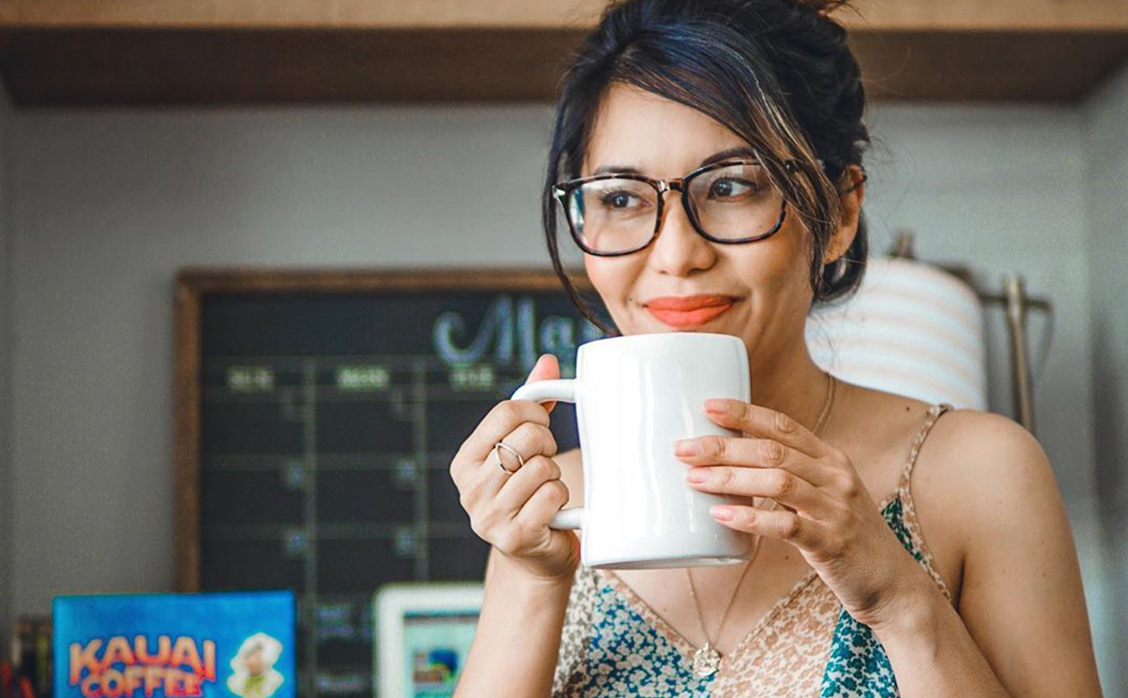 a woman drinks coffee out of a white mug. A box of kauai coffee is visible on the desk behind her.